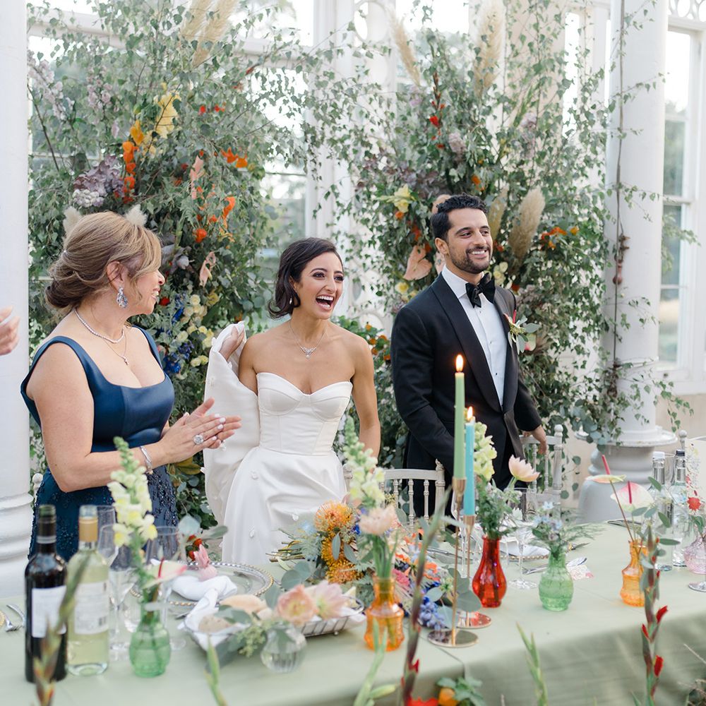 Bride & groom stand in front of brightly coloured floral installation complete with small colourful vases on pastel green tablecloth 