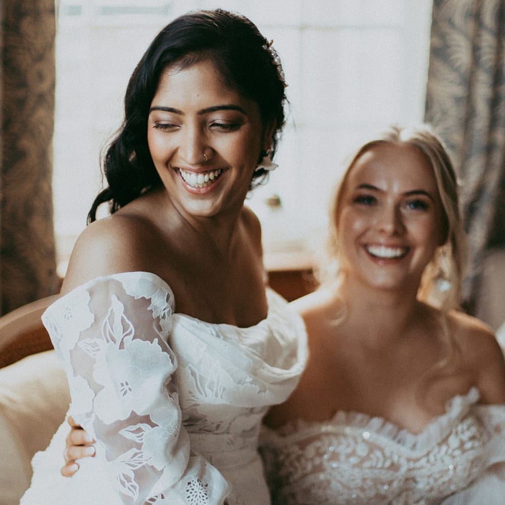 Bride in lace and diamond detailed off shoulder wedding dress sitting with bride in off shoulder long sleeve wedding dress with lace floral detailing