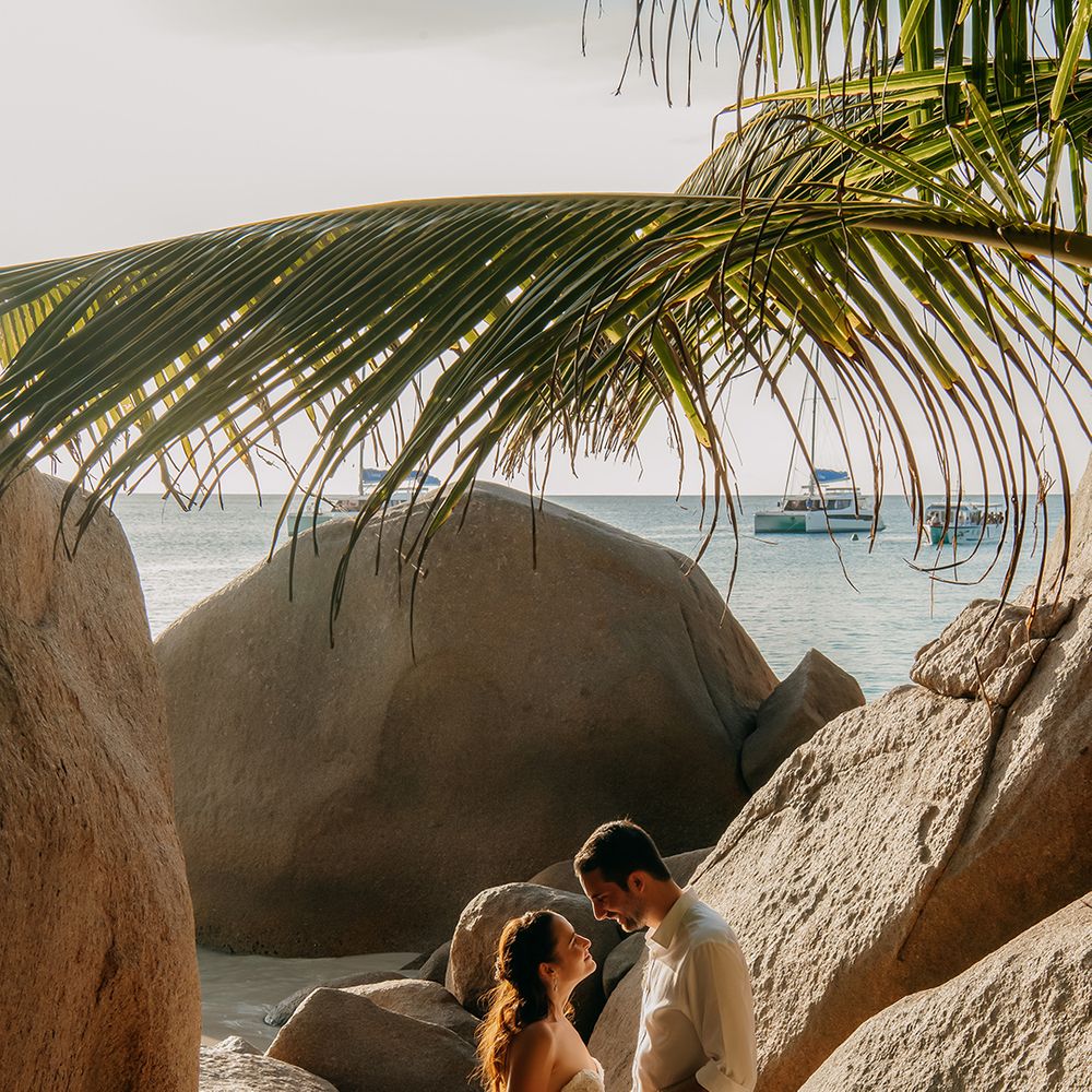 Bride in off the shoulder lace wedding dress with sweetheart neckline and lace floral details and groom in crisp white shirt, light coloured chinos, tan leather belt and white trainers holding hands on a beach in Seychelles