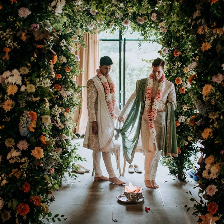 Grooms stand between colourful floral arch during barn wedding 