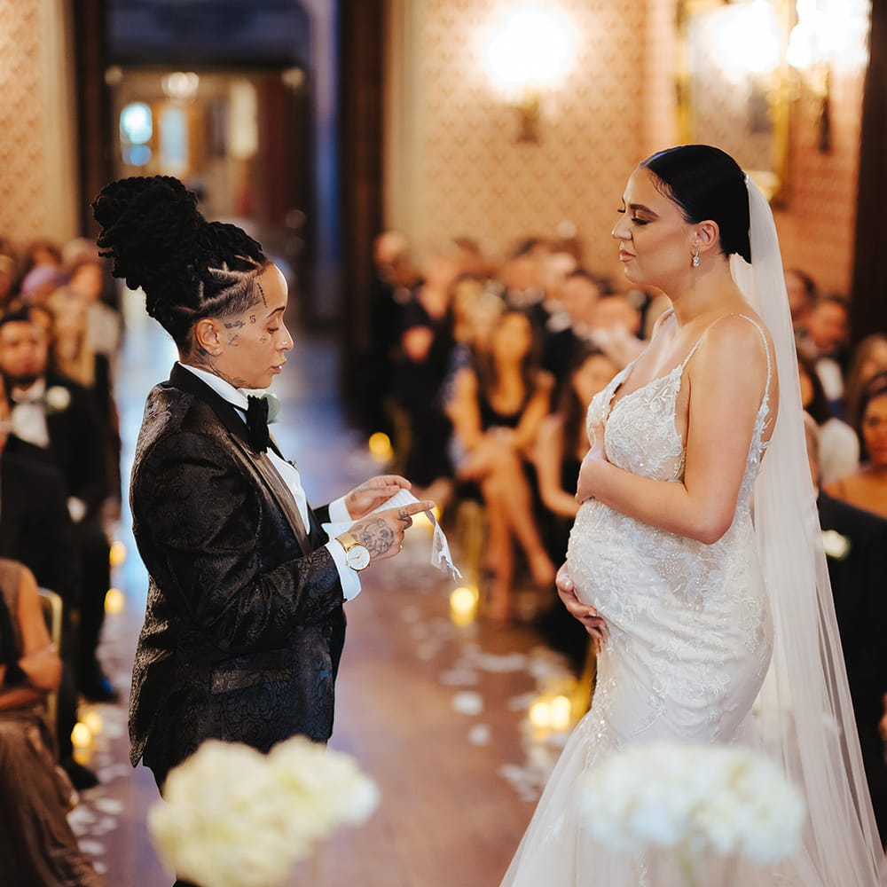 Bride in lace sleeveless v neck Enzoani wedding dress with tulle finish and church length veil holding baby bump listening to bride saying their vows in black snakeskin print tuxedo with black bowtie and white rose boutonniere