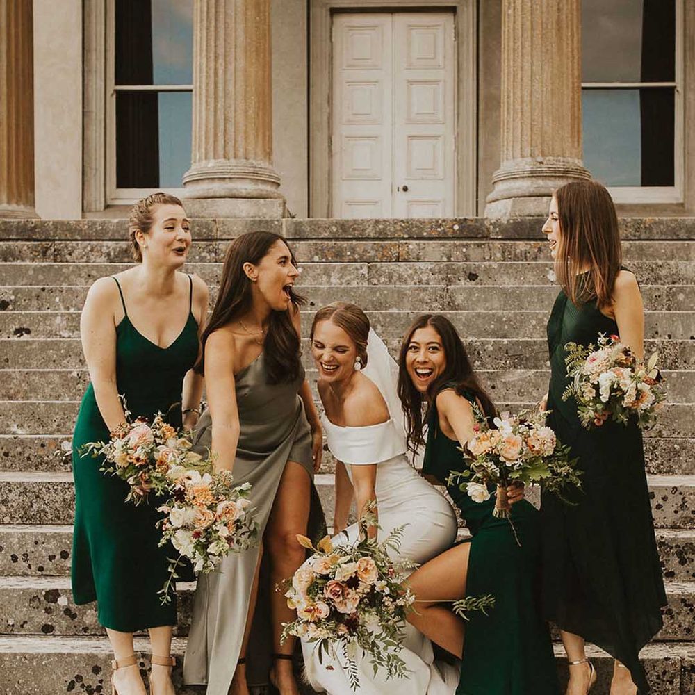 Bride in off the shoulder bodycon wedding dress standing on the steps of The Grange Hampshire with bridesmaids in mismatched green bridesmaid dresses 