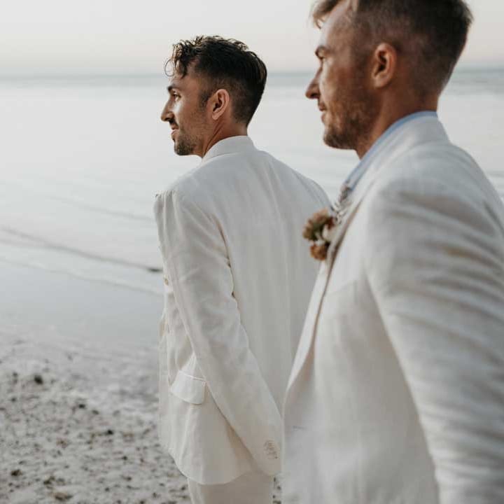 Grooms in complimenting double breasted white wedding suits holding hands doing couples photoshoot on the beach in Holbox 
