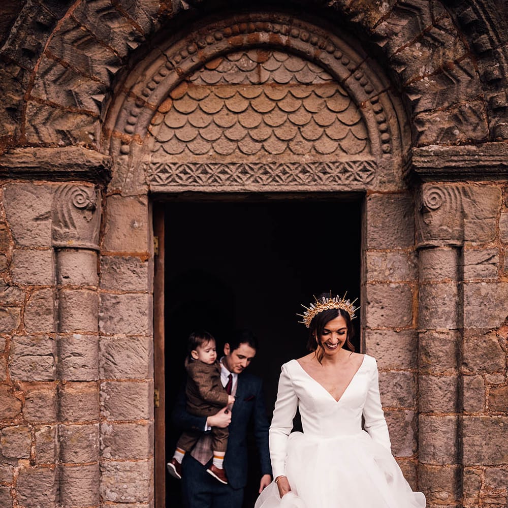 Bride in a ruffle gown and crown walking out of their wedding ceremony with the groom carrying their son 