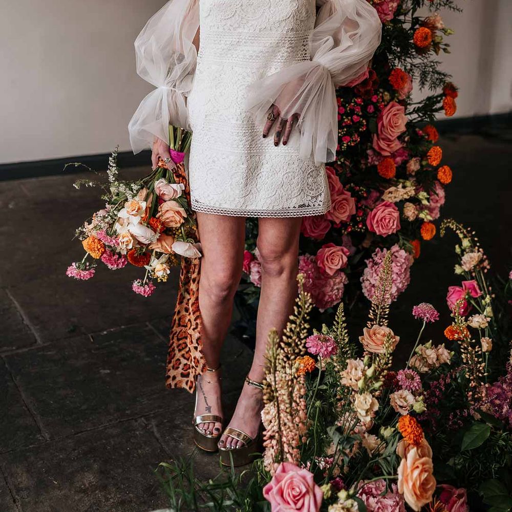 Bride wearing 70s inspired boatneck short lace wedding dress with detachable tulle puff sleeves and platform gold wedding heels standing by circular floral arch with vibrant flowers and foliage attached