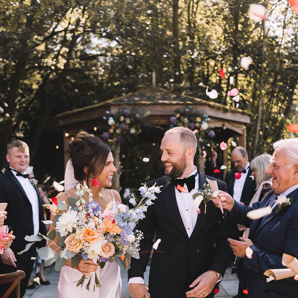 Bride in satin slip wedding dress walking with groom out to colourful confetti exit 