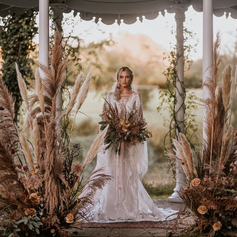 The bride stands underneath the band stand holding an epic pampas grass bouquet, surrounded by pampas grass florals