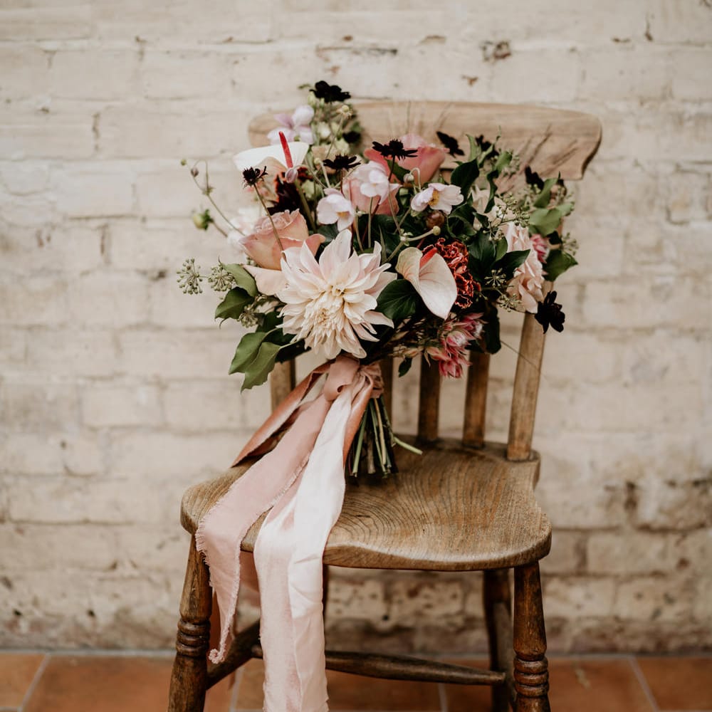 Blush pink and green wedding bouquet with anthuriums, roses and dahlias tied with ribbon sitting on a wooden chair 