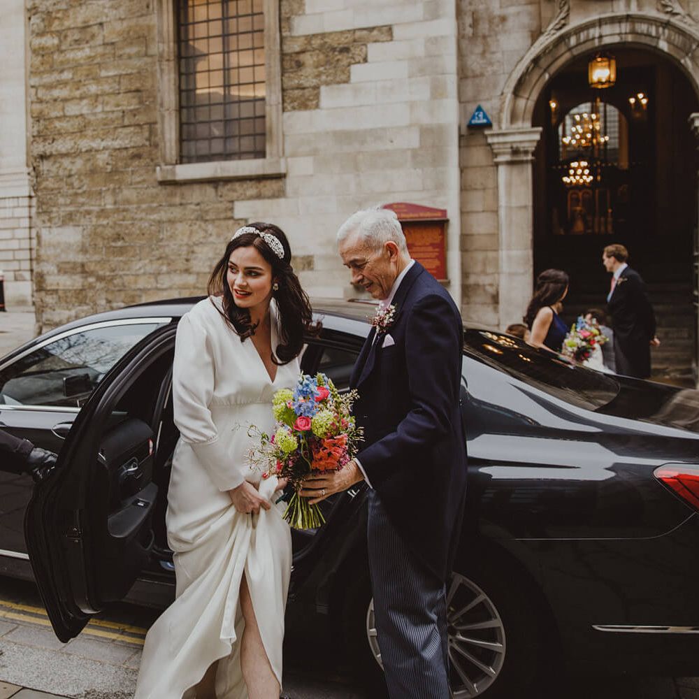 Bride stepping out of wedding car wearing front split, long sleeve wedding dress holding bright poppies for London church wedding