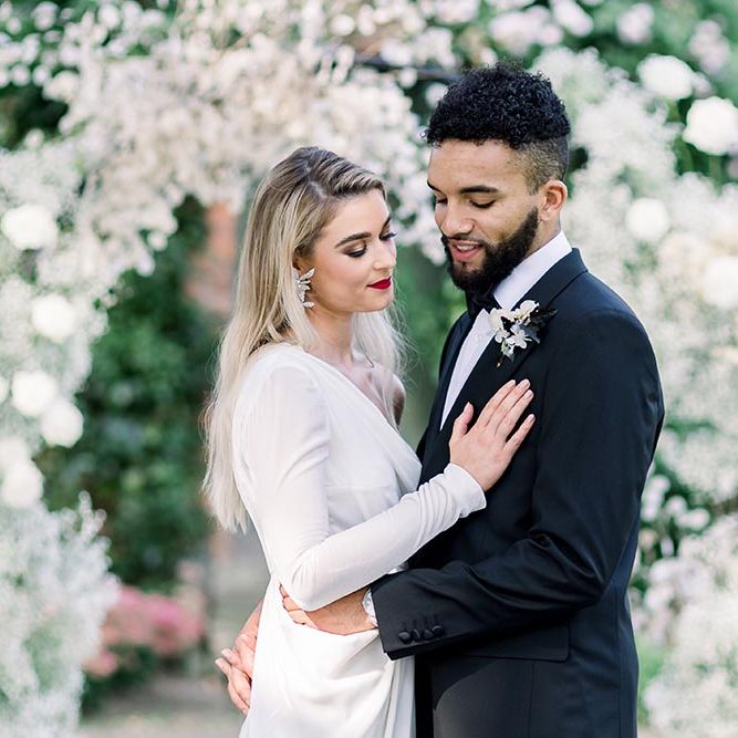 Groom in a tuxedo embracing his bride in a one shoulder long sleeve wedding dress with wrap skirt 