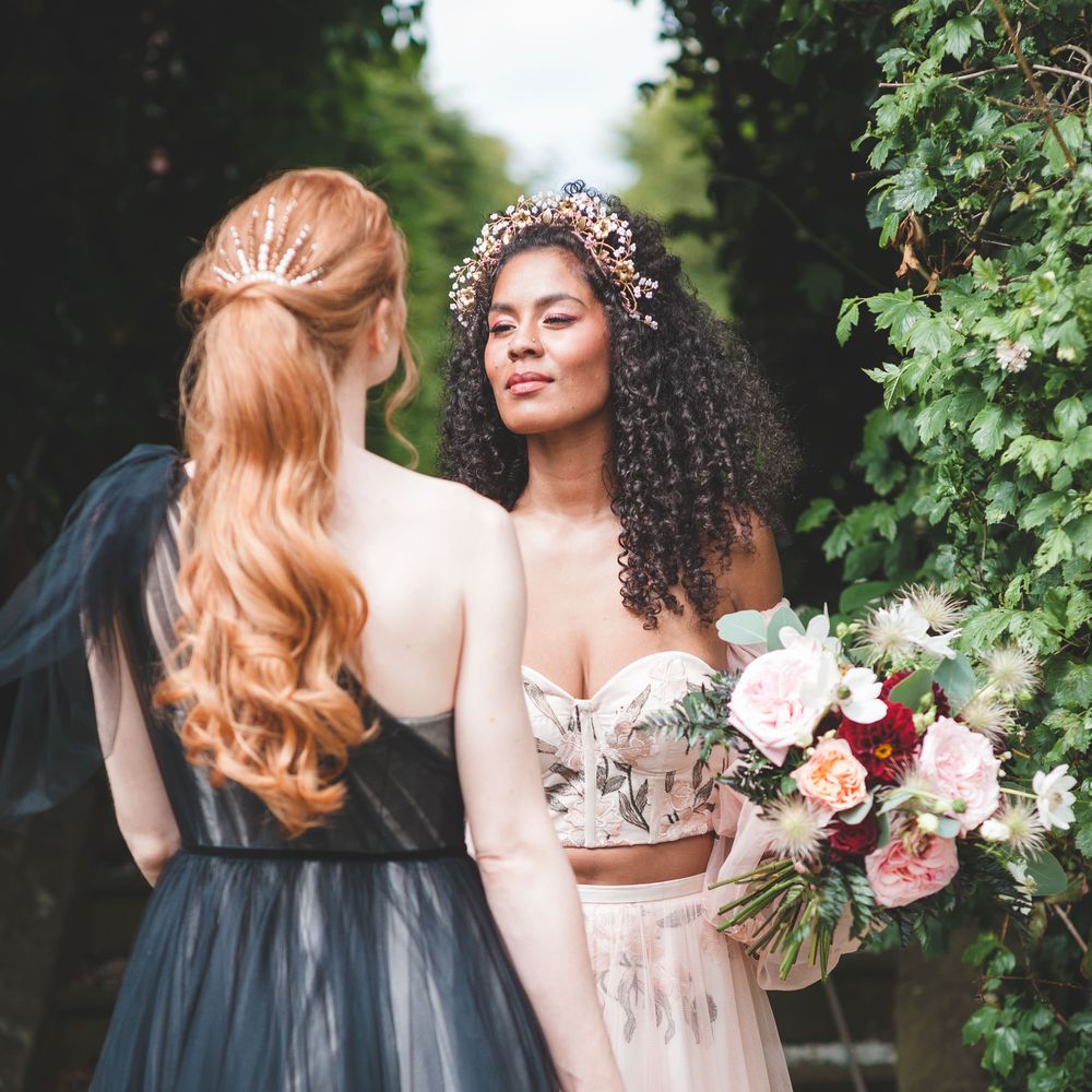 Black bride with long naturally curly hair wearing a bridal headdress in an embroidered bodice and tulle skirt looking at her bride to be