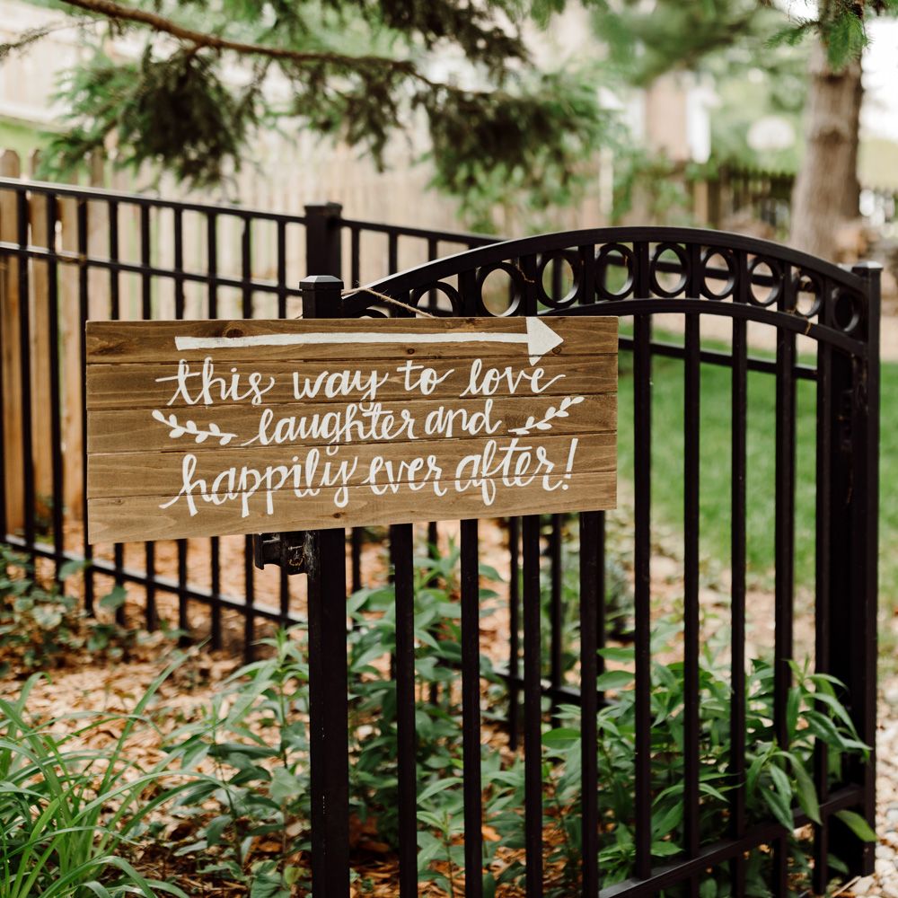 Wooden sign with white painted words reading 'this way to love laughter and happily ever after' hanging on black gate at backyard wedding