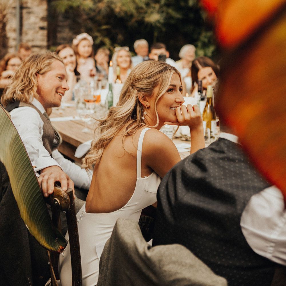 Smiling bride sits next to smiling groom at wooden wedding breakfast table in glasshouse at Anran Devon as they watch speeches 