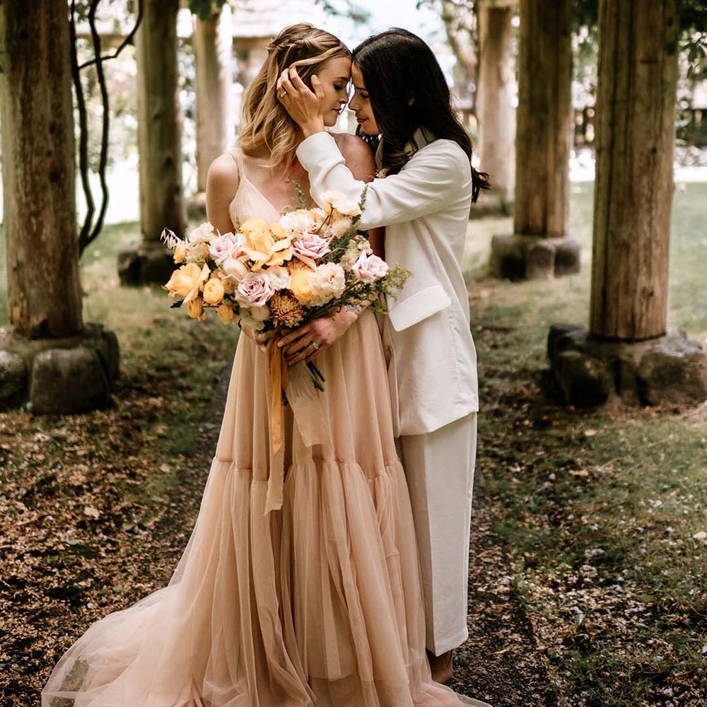 Bride in a white suit embracing her brides face in a blush pink tulle wedding dress as she holds a peeled back rose wedding bouquet 