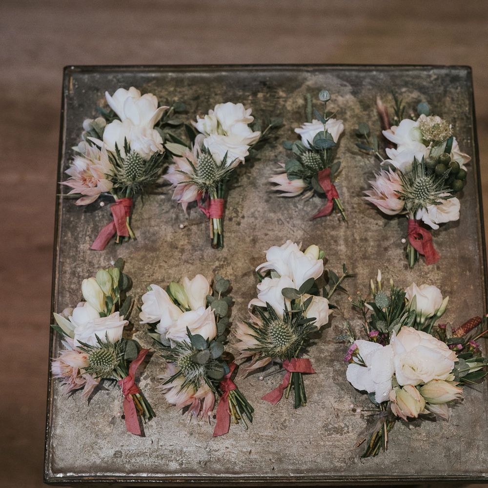 Eight protea, rose and thistle buttonholes tied with red ribbon at Tythe Barn wedding with barn wedding flowers