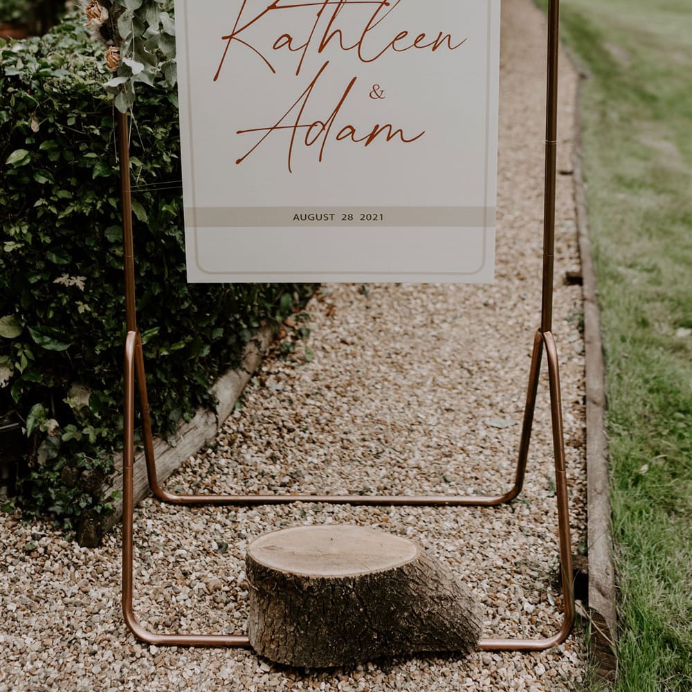 Copper frame wedding sign reading 'Welcome to the wedding of Kathleen and Adam August 28 2021' with foliage decor