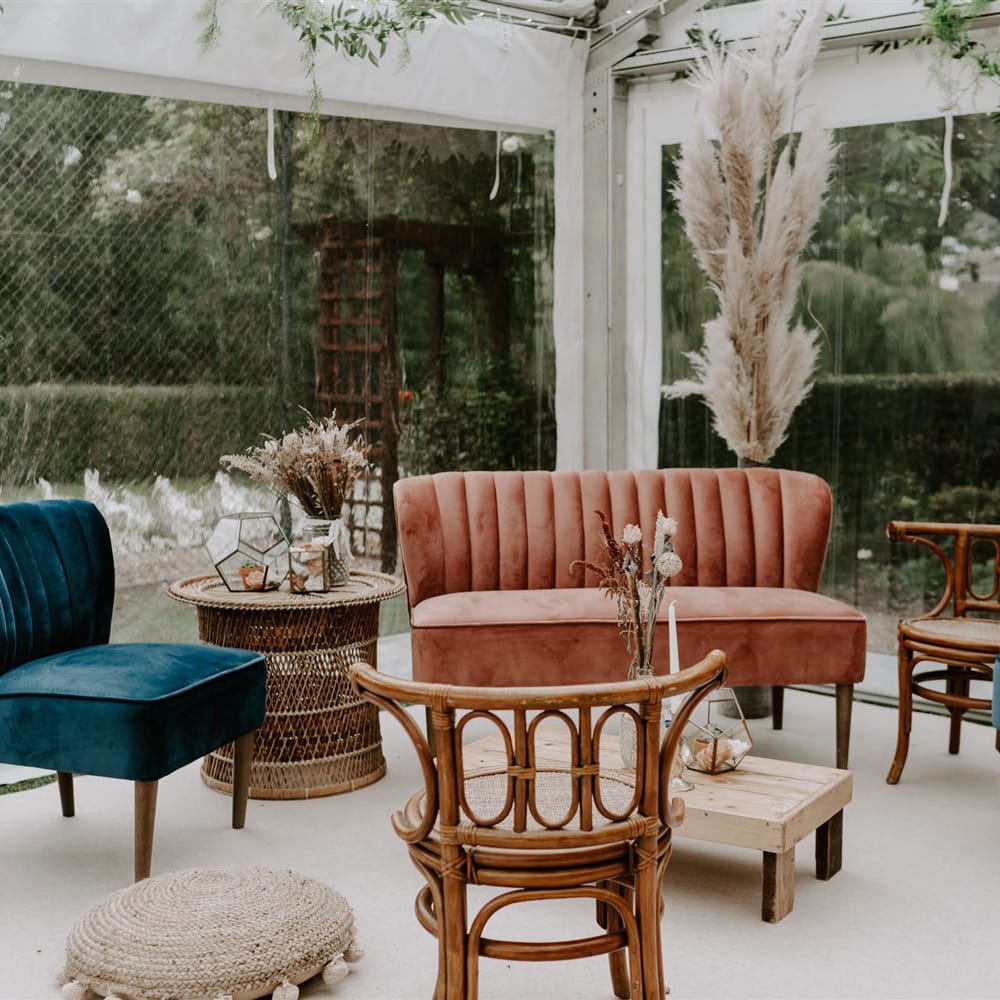 Pink velvet sofa and two blue velvet occasion chairs with various wooden furniture in corner of marquee at wedding reception with garlands and pampas grass for garden wedding