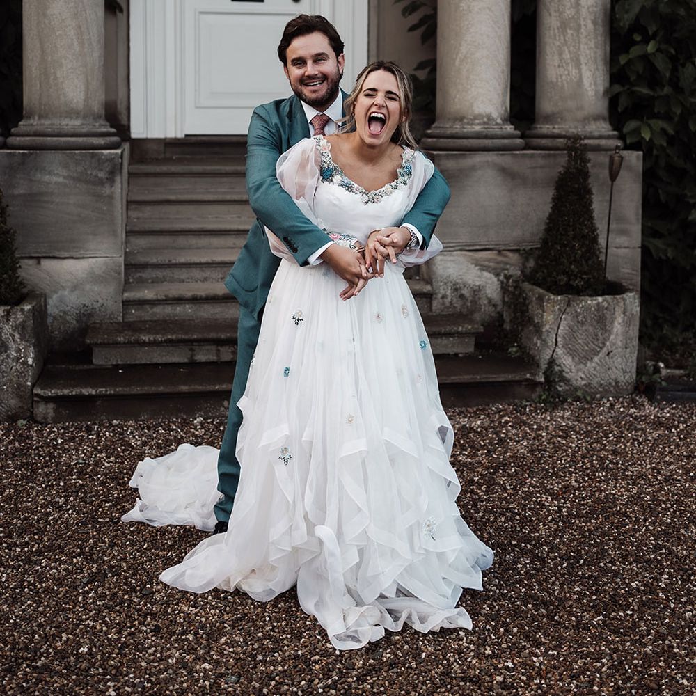Laughing bride in long sleeve wedding dress with floral embroidery is hugged by groom in blue suit outside the front of Iscoyd Park