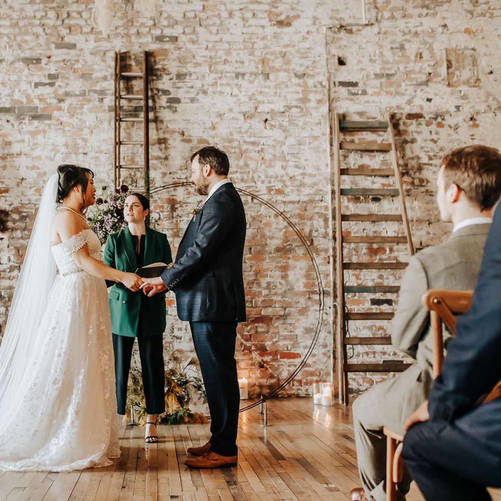 Bride in off the shoulder wedding dress, veil and pearl necklace stands holding hands with groom in blue suit and brown shoes during non denominational wedding ceremony