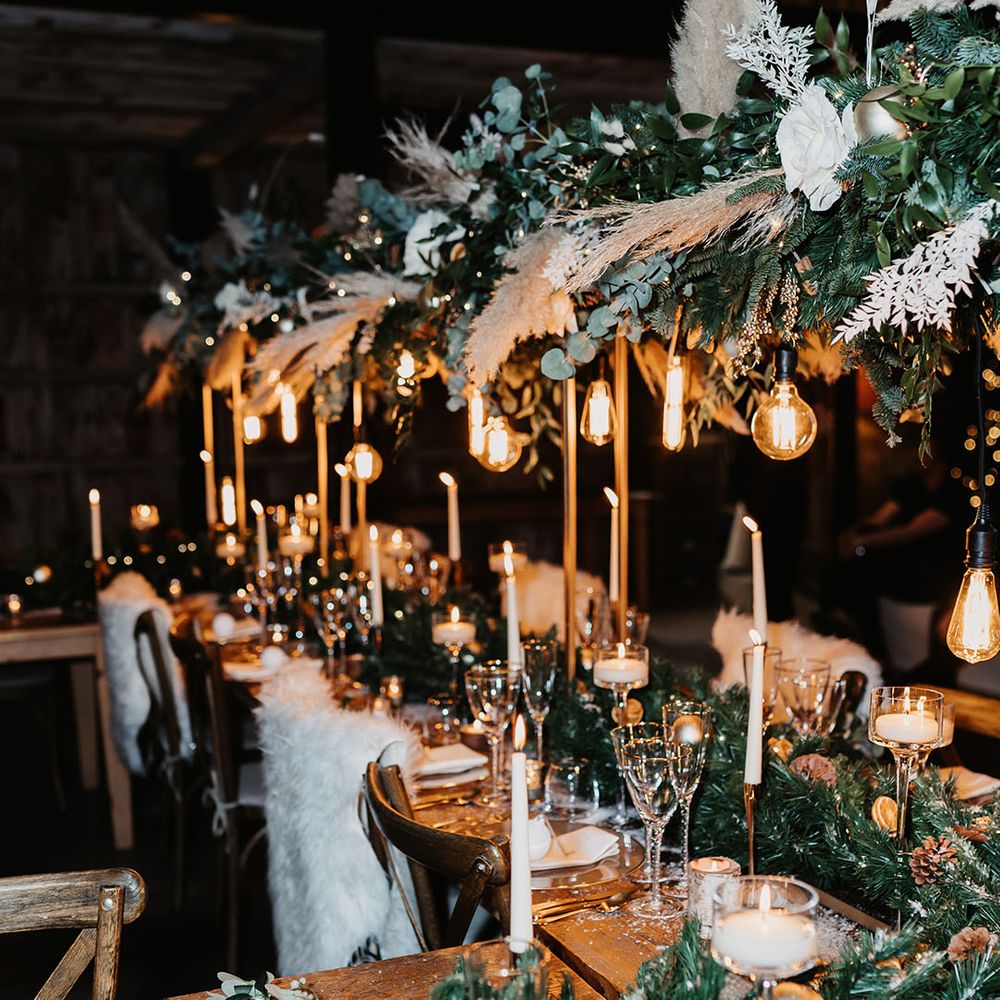 White fur throws on backs of chairs with pinecone runner, white candles, gold rimmed glasses and exposed lightbulbs