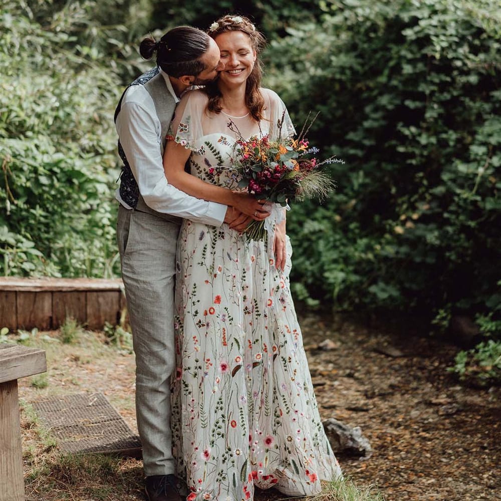 Groom in grey trousers and waistocat kissesthe bride on the cheek who wears a floral embroidered wedding dress with colourful bouquet 