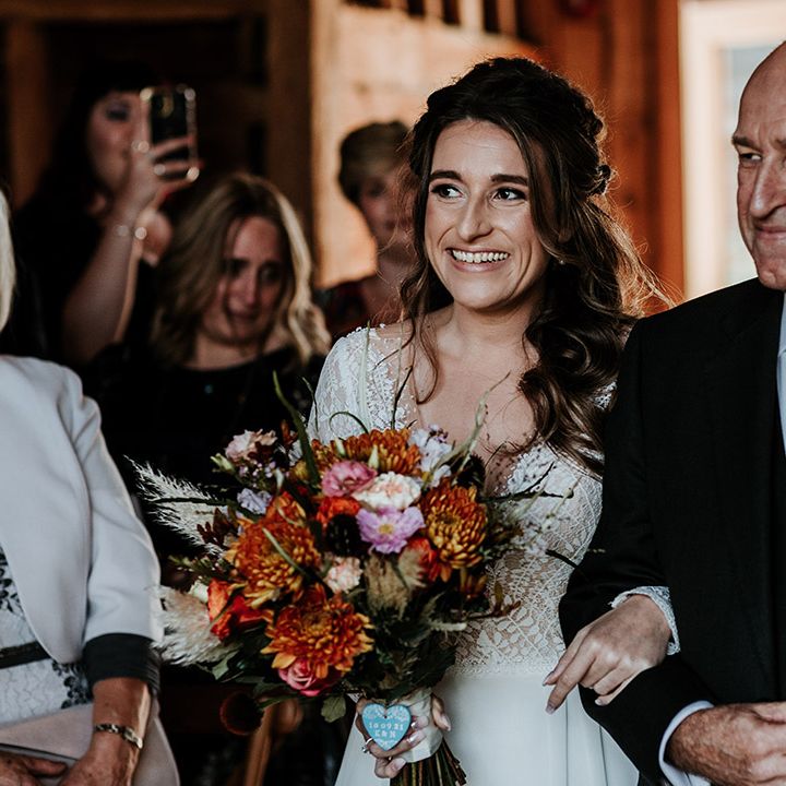 Bride with curled half up half down hair holds orange and red wedding bouquet is walked down the aisle by father of the bride in emerald green tie