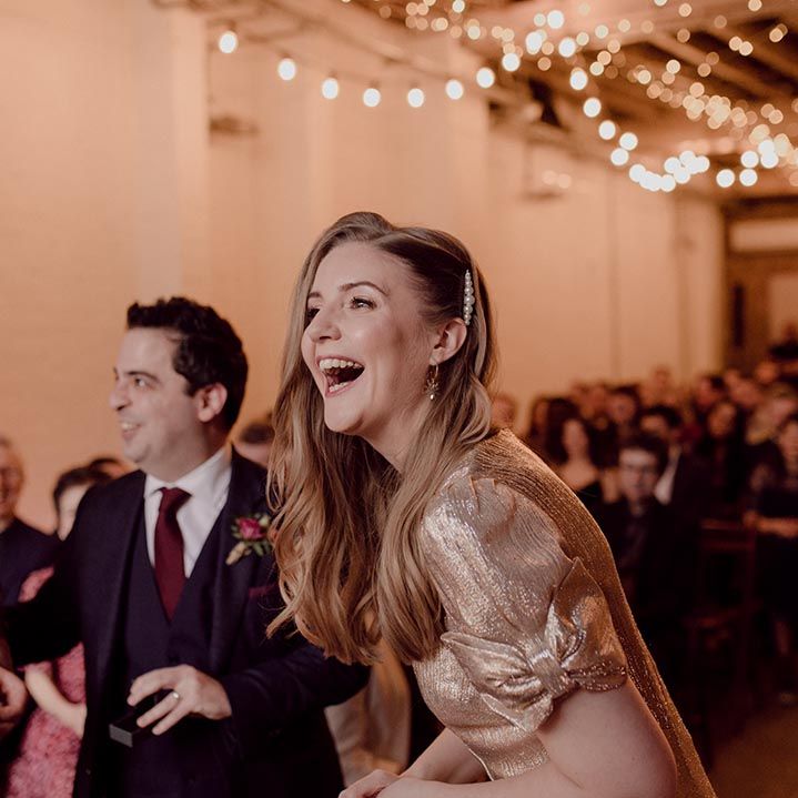 Bride in sparkly gold wedding dress with bow sleeves and pearl hair clip smiles standing at the aisle with the groom in a velvet red suit jacket