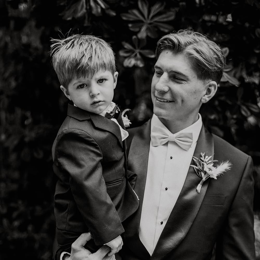Groom in black tie with pale blue bow tie holds son in black tie for wedding 