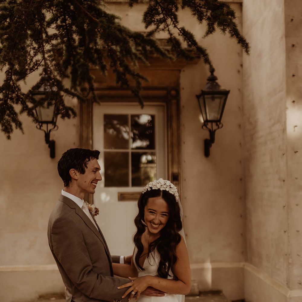 Groom in neutral wedding suit holds the bride around her waist while she wears an Alexandra Grecco wedding dress and flower bridal headband