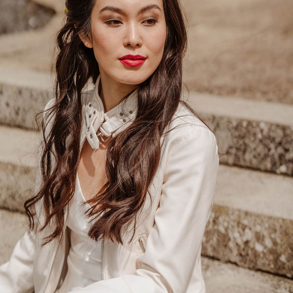 East Asian bride with long wavy hair wearing a flower in har hair, red lipstick and a white leather jacket 