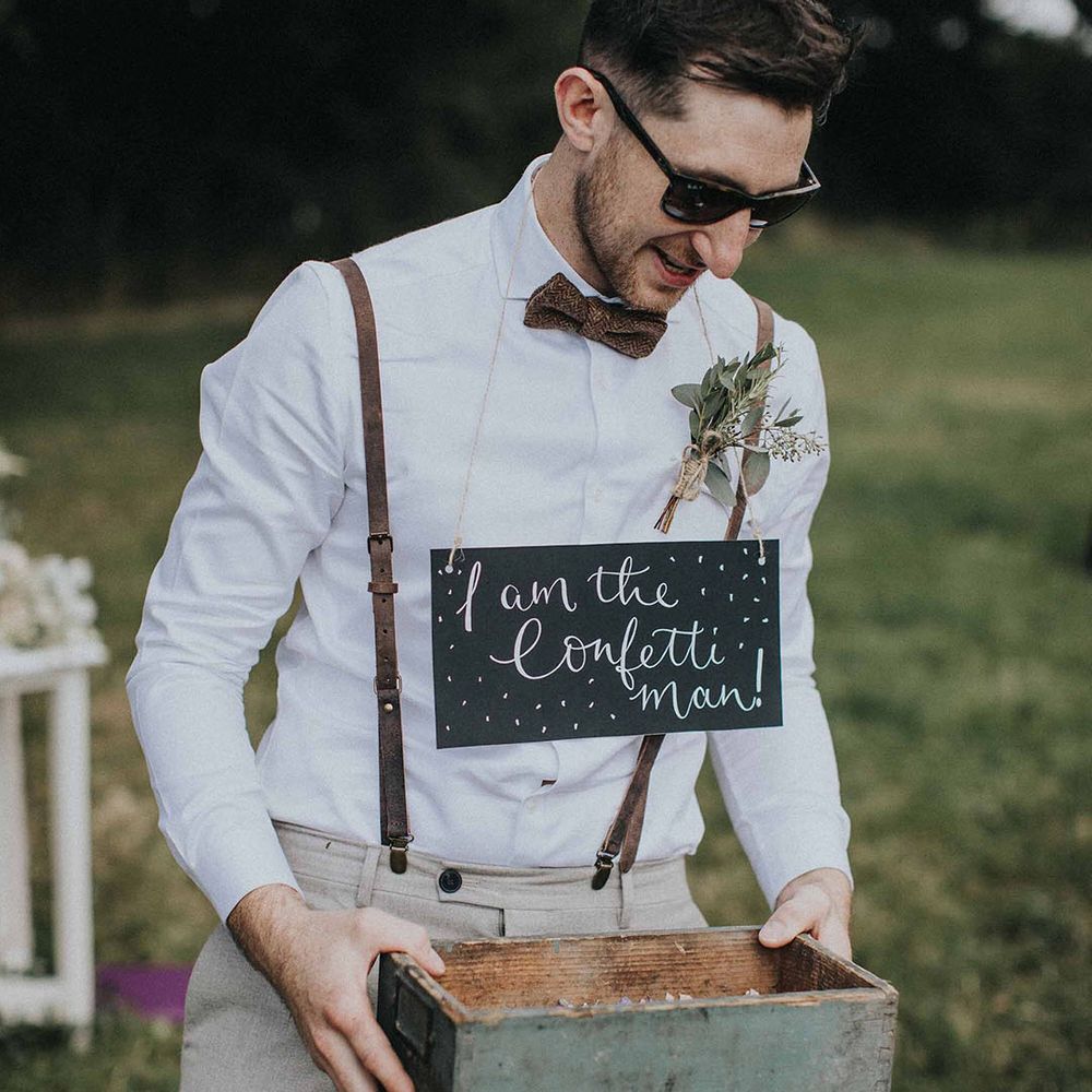 Wedding guest holding a box of confetti with a chalkboard sign reading 'I'm the confetti man' in sunglasses and braces for his informal role in the wedding
