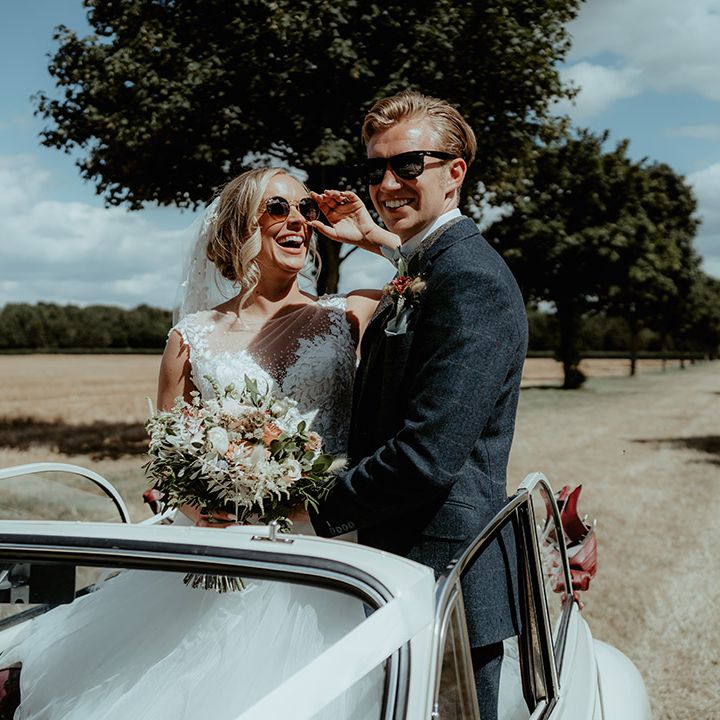 Bride and groom stand up in their cream wedding car with sunglasses on 
