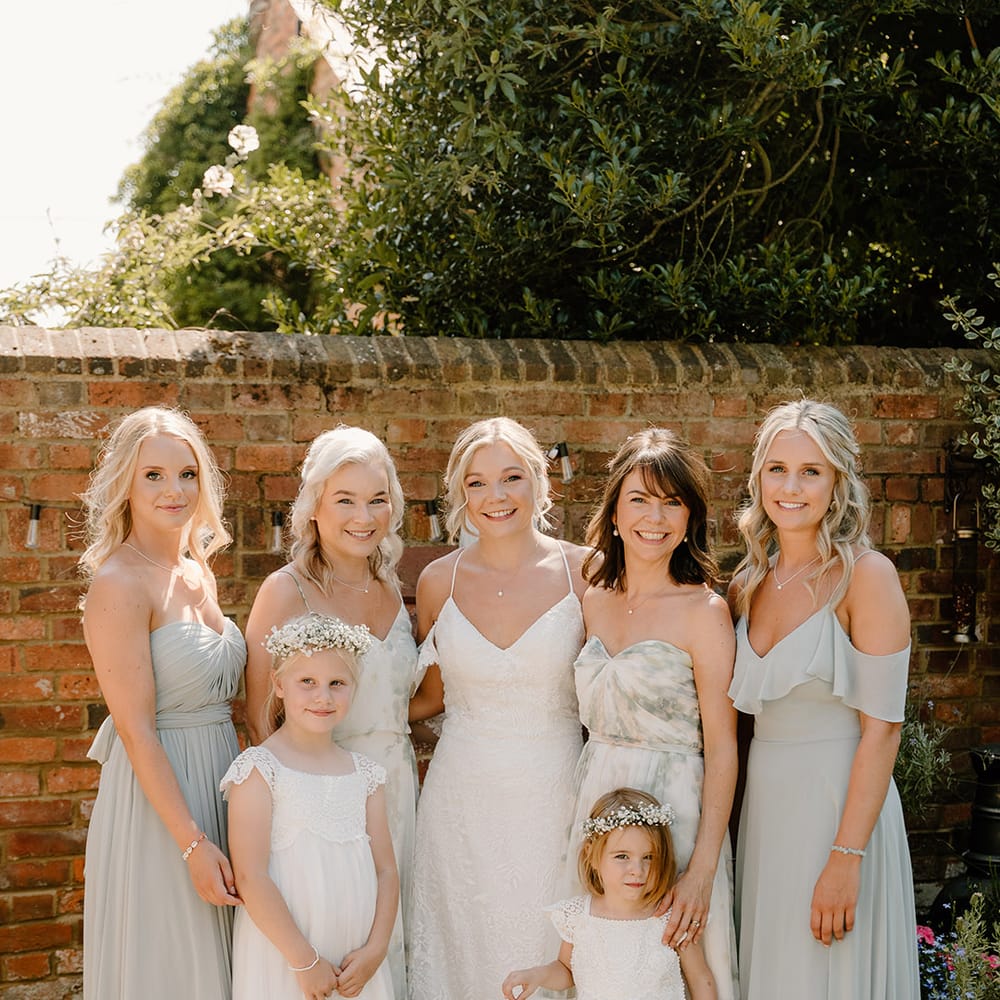 Bridesmaids wear different styled sage green dresses whilst stood beside bride in lace fitted wedding dress