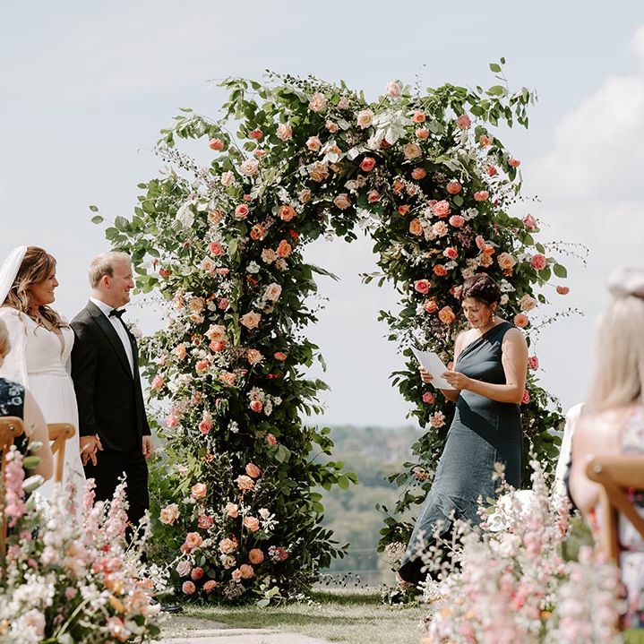 Bridesmaid in one shoulder green bridesmaid dress reads in front of floral archway filled with orange and pink roses surrounded by green foliage