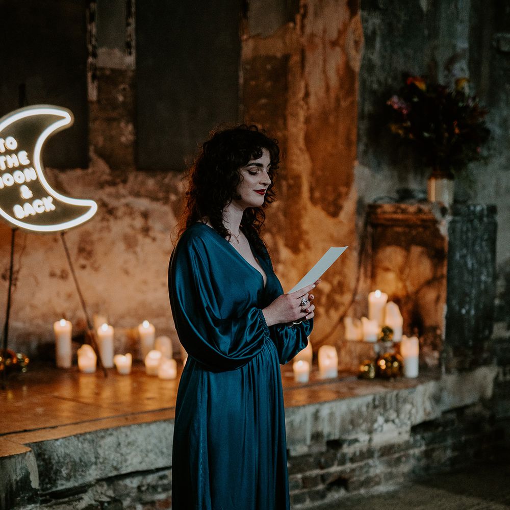 Bridesmaid in satin green dress doing a wedding reading at The Asylum Chapel surrounded by candles and a moon neon sign