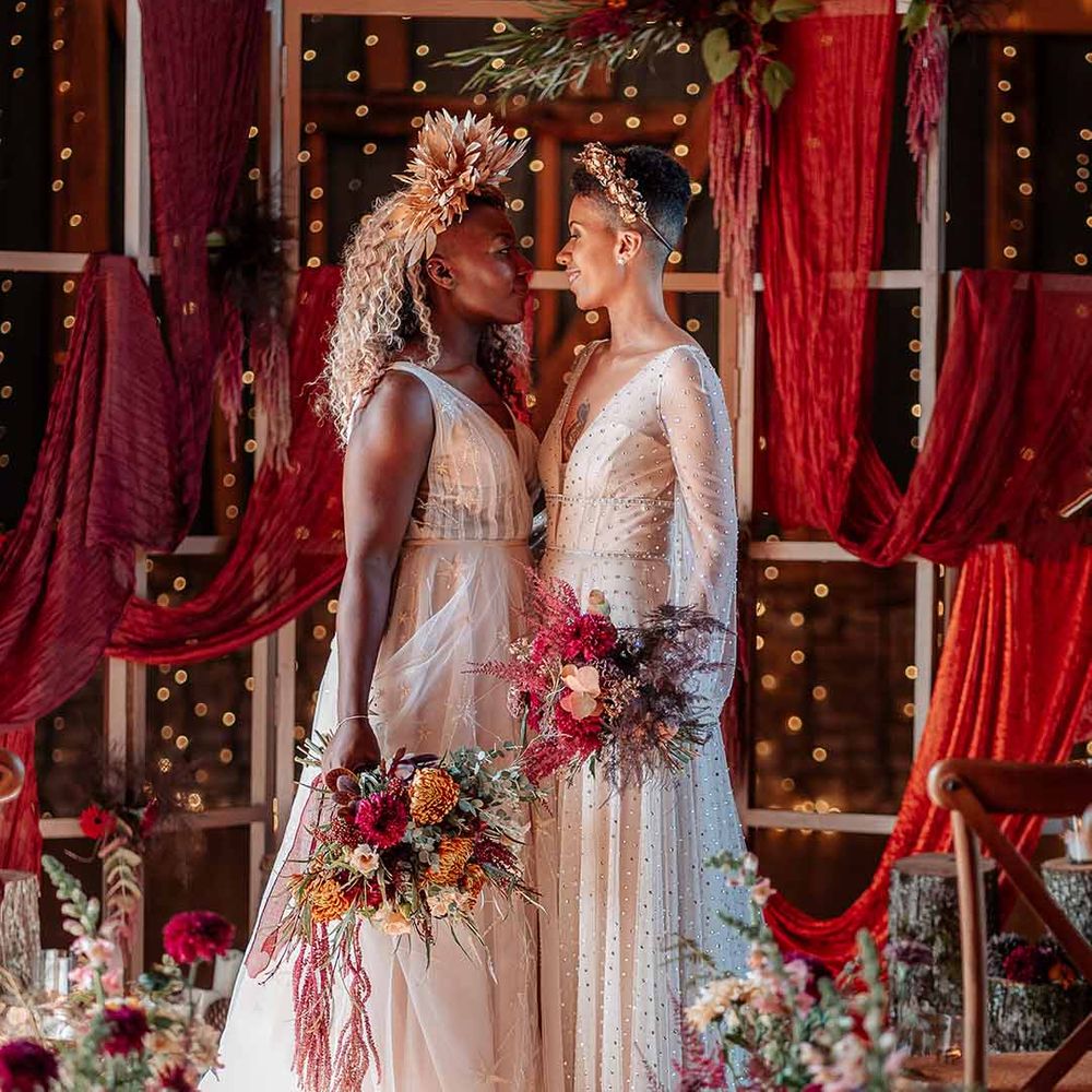 Bride in crystal embellished gown and gold headdress and sleeveless floaty peach gown and gold leaf crown standing with red wedding drapes, fairy lights and autumnal dried flower arrangements