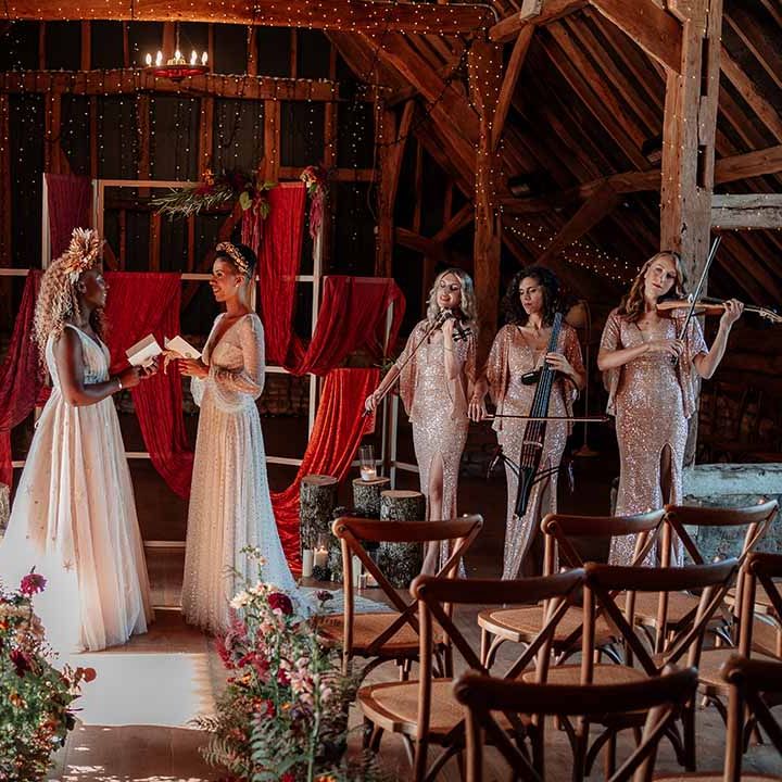 Brides reading their vows in front of square wooden decor with red and burnt orange wedding drapes, foliage and autumnal dried flower decor, fairy lights, festoon chandelier and string trio