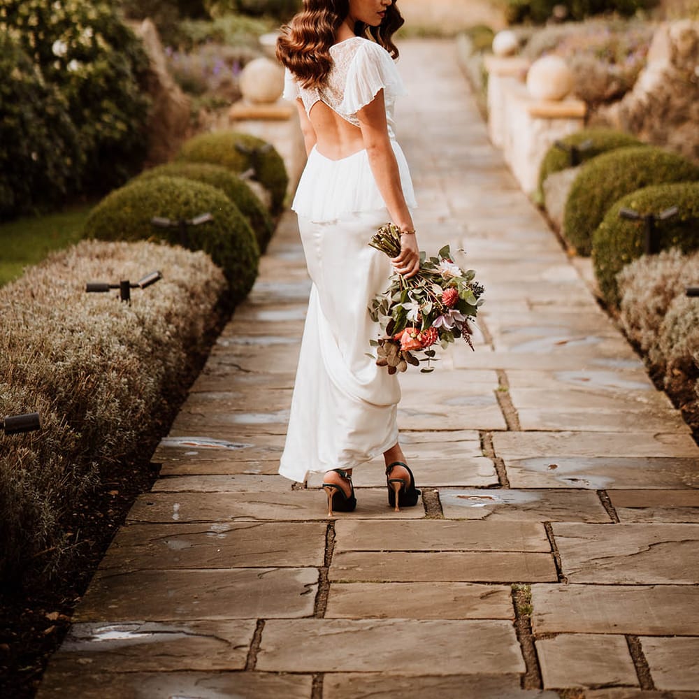 Bride holding wedding bouquet with white and pink carnations, garden roses, eucalyptus, baby’s breath and dried flowers wearing satin open back wedding dress with button details and butterfly sleeves, ruffled and layer on the back and strappy black high heels