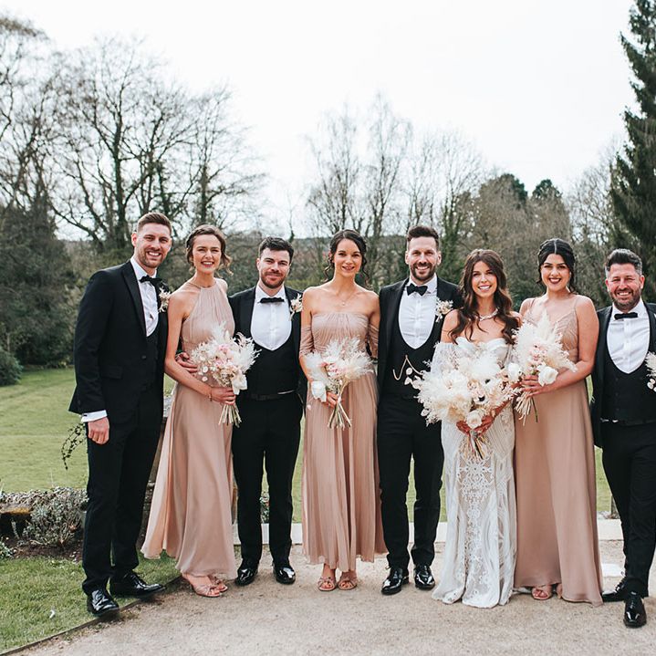Wedding party shot of the bridesmaids in neutral pink dresses in different styles with the groomsmen and groom in black tuxedos and the bride in a fitted lace wedding dress 