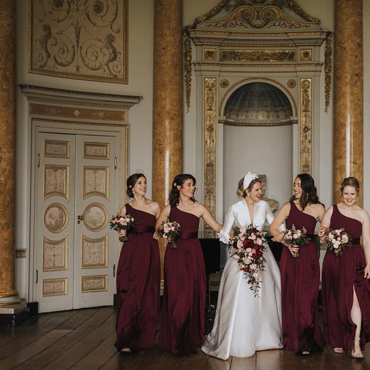 Bridesmaids in one shoulder burgundy dresses walk with the bride in a traditional wedding dress holding their matching pink and red bouquets 