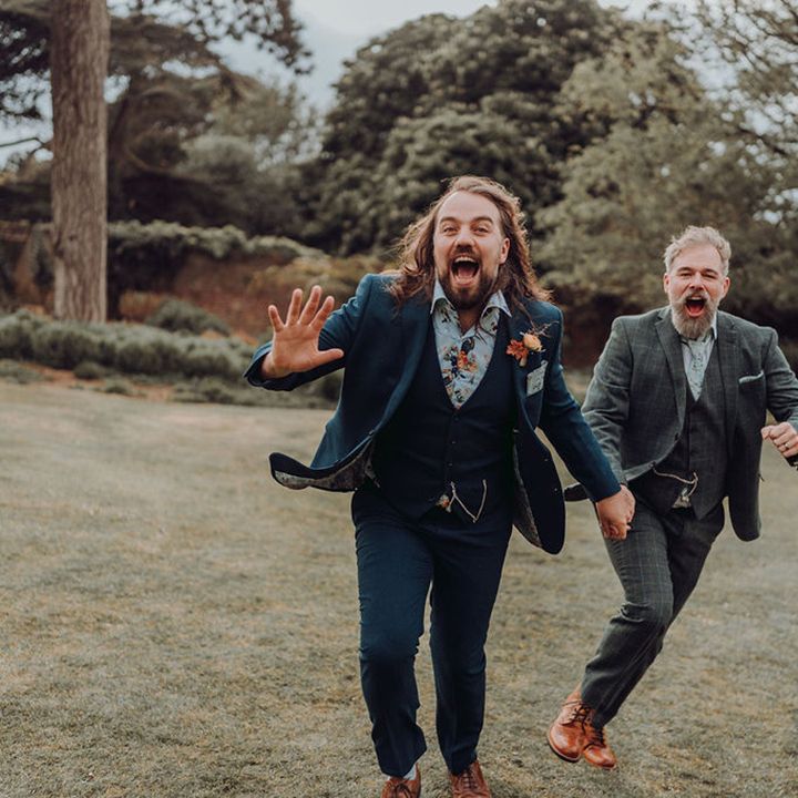 Groom in blue suit and floral shirt hold hands alongside his groom whilst running with one another outdoors 