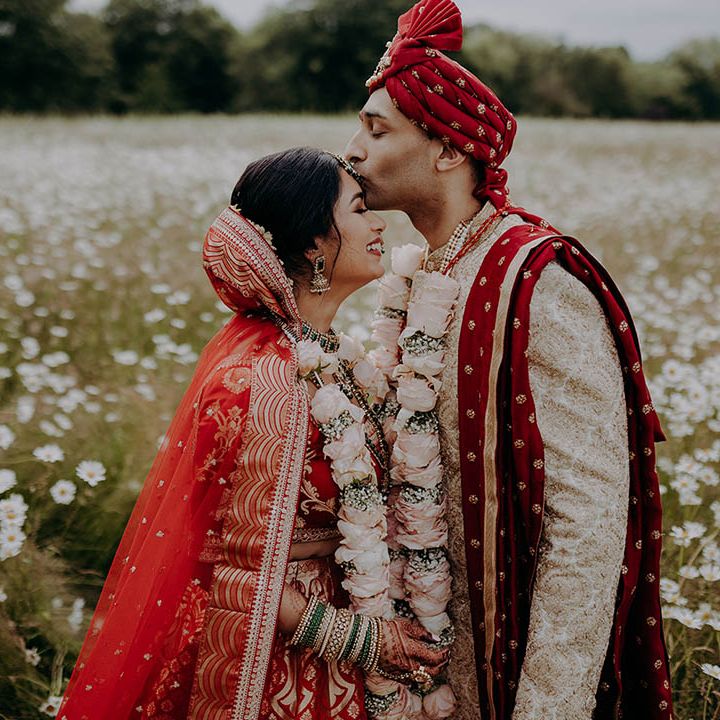 Groom in red, white and gold sherwani kisses his brides forehead during outdoor couples portraits 