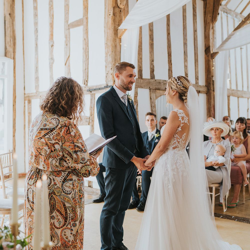 Groom in three piece navy suit stars facing the bride holding her hands in an open back illusion wedding dress for rustic barn wedding 