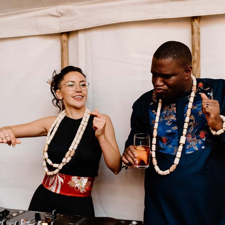 Bride in black bridal jumpsuit with red and gold Chinese fabric belt dancing with groom in traditional Nigerian wedding outfit both wearing white beads around their neck at Broadfield Court