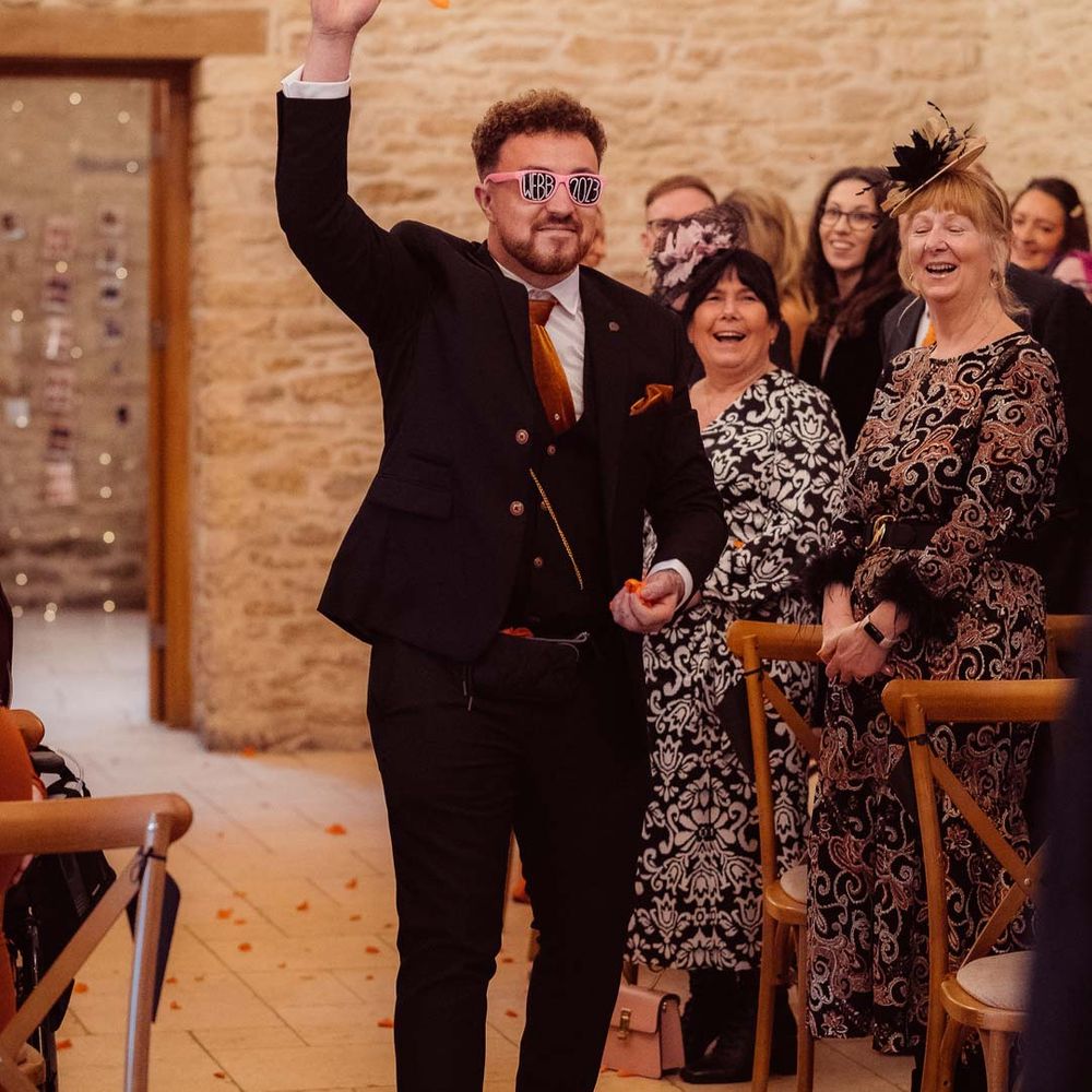 Wedding guest wearing dark suit with burnt orange wedding tie and pocket square walking down the aisle at Kingscote Barn Tetbury 