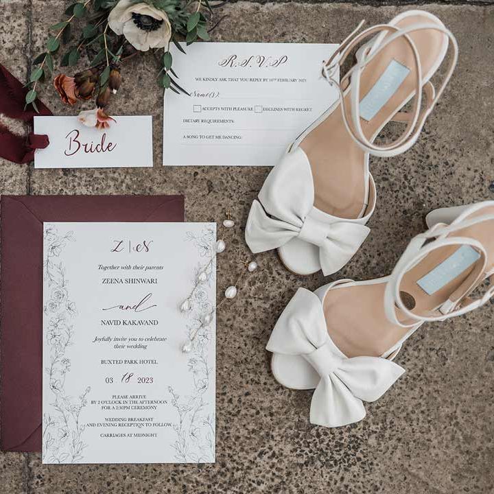 Minimalist white and maroon wedding stationery with fine line drawings and calligraphy surrounded by dried flowers, maroon ribbon and white open toed wedding heels