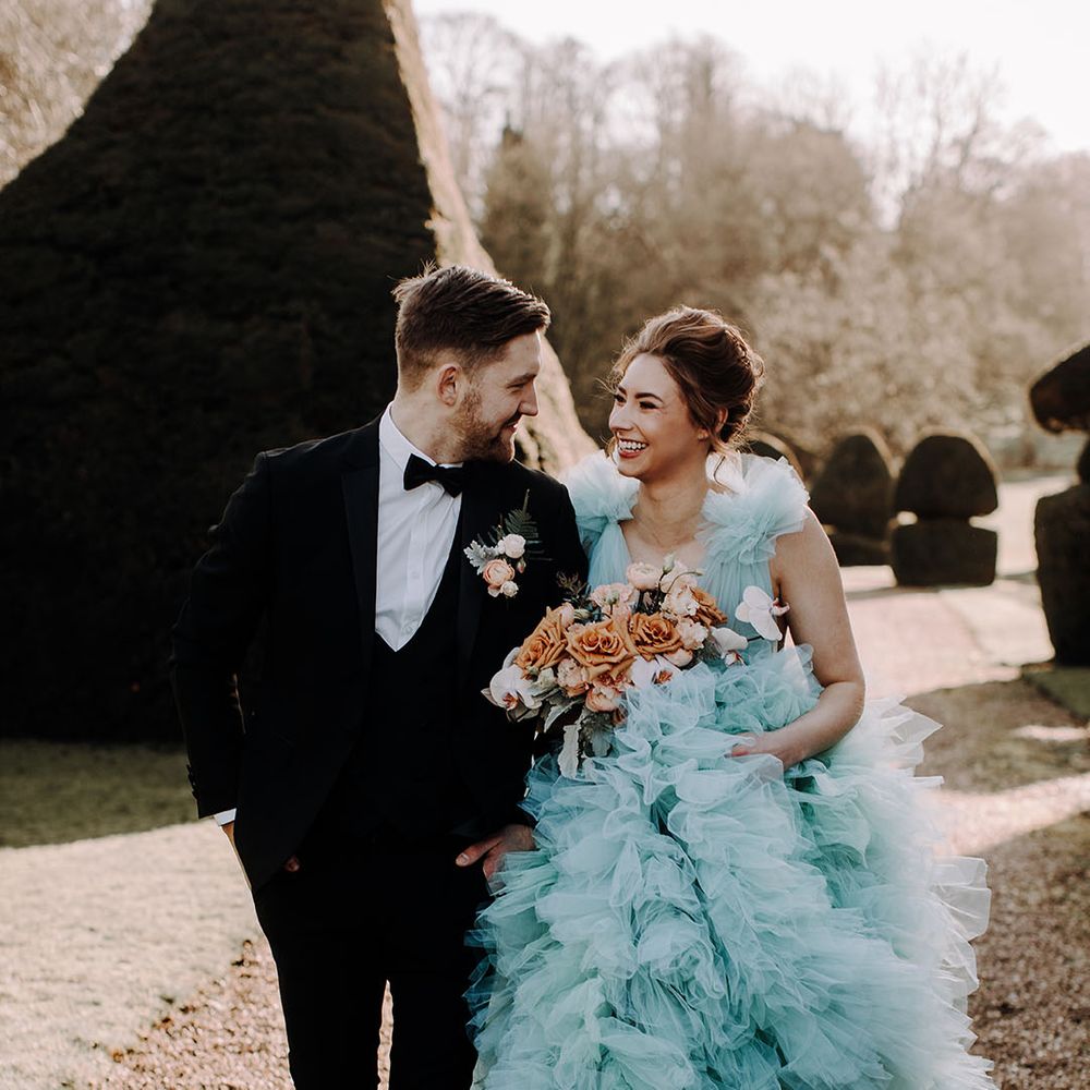 Bride wearing ruffle tulle light blue wedding dress with orange rose bouquet and groom in black tuxedo 