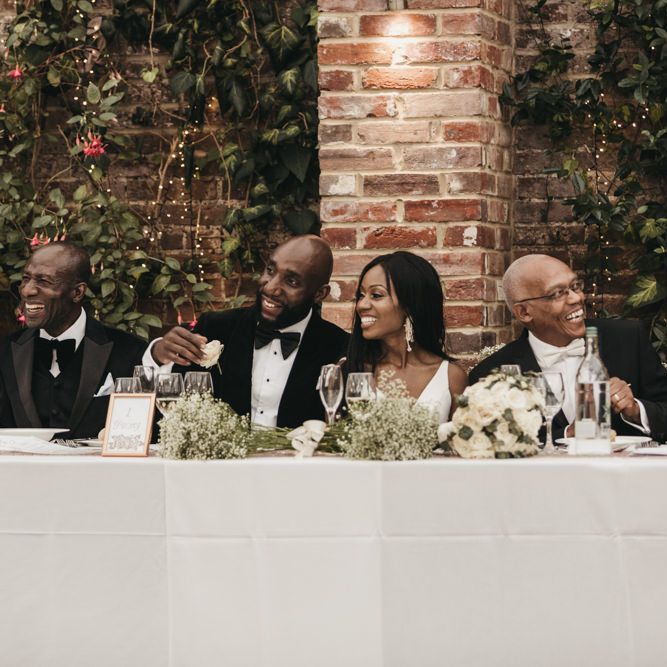 Black bride and groom sitting at the top table decorated with white and green flowers with their parents 