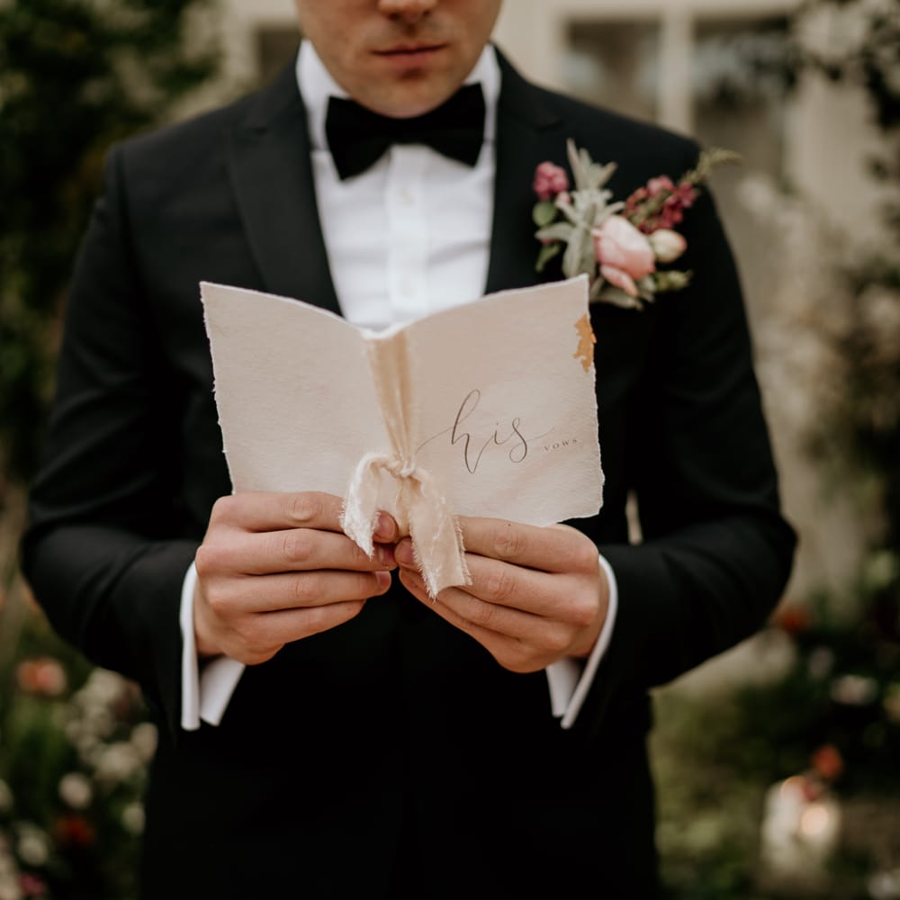 Groom in a black tuxedo with bow tie holding his vow book tied with ribbon