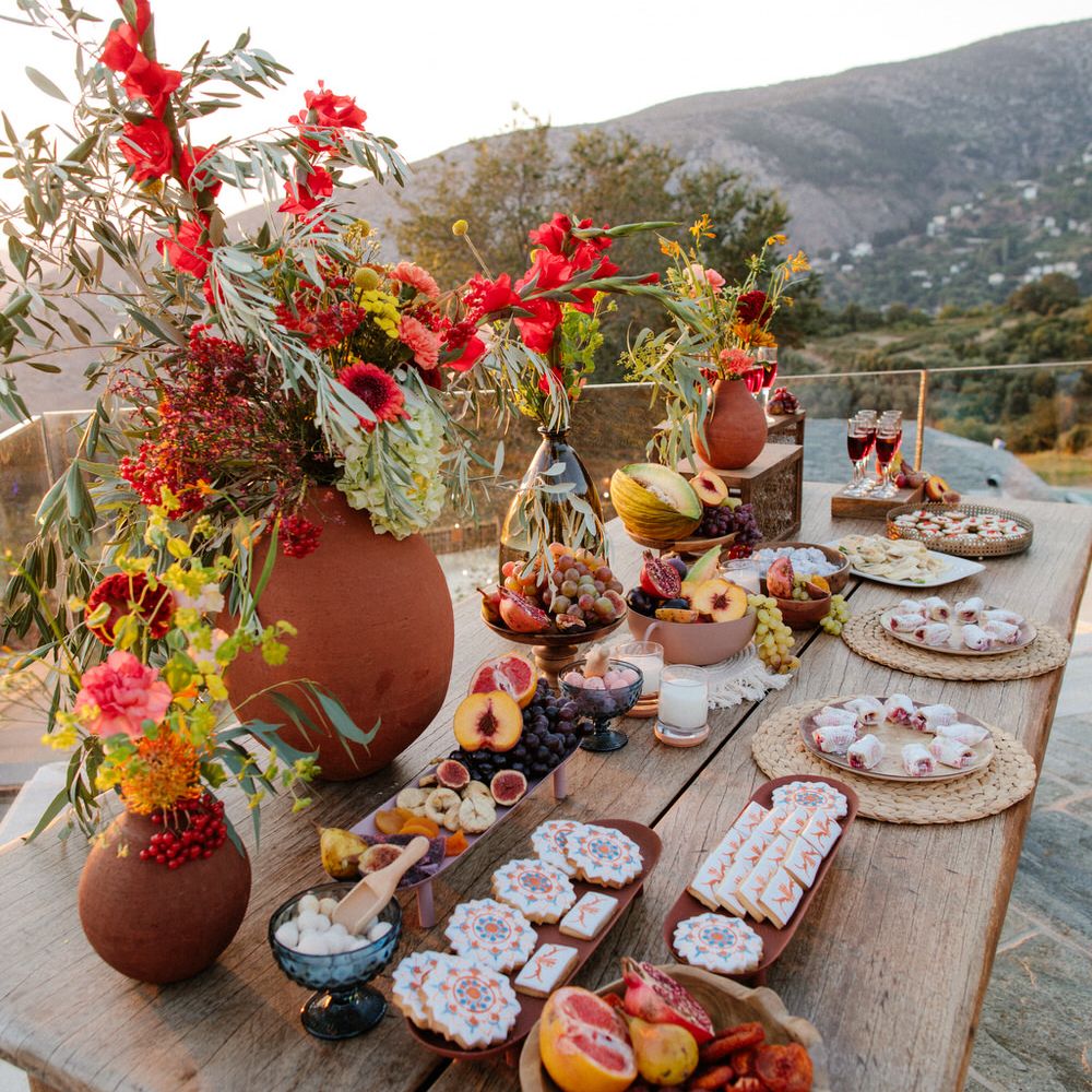 A grazing table full of local Greece produce including cookies, fruit and desserts, and terracotta vases with wildflowers